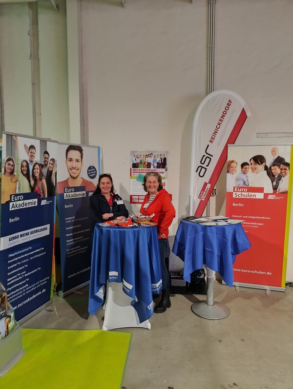 Euro Akademie und Euro-Schulen Berlin informieren Eltern bei „Komm auf Tour“ Zwei Frauen, eine in dunkelblau und eine in rot gekleidet, stehen am Messestand der Euro Akademie und Euro-Schulen Berlin und lächeln in die Kamera.