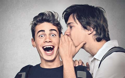 Symbolbild für positive Neuigkeiten Ein junger Mann flüstert einem anderen jungen Mann etwas ins Ohr und verdeckt dabei seine Lippen mit seiner Hand. Der andere Mann hat den Mund lächelnd weit geöffnet und die Augenbrauen weit nach oben gezogen. Er scheint eine positive, überraschende Botschaft zu erhalten.