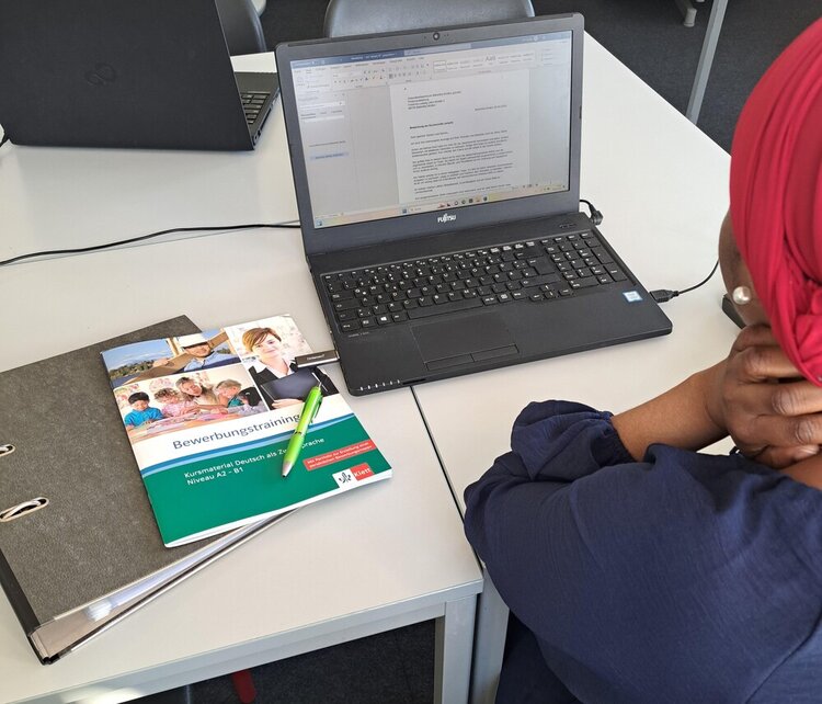 Erfolgreicher Start des Bewerbungscoachings für Migranten Eine Frau, von der man nur die Rückansicht sieht und die ein rotes Kopftuch und einen Perlenohrring trägt, sitzt an einem Tisch vor einem Laptop, auf dem man ein Bewerbungsschreiben erkennen kann. Im Laptop steckt ein USB-Stick. Neben dem Laptop liegt ein geschlossener Ordner und darauf ein Kuli sowie ein DIN A4-Heft aus dem Klett-Verlag mit dem Titel "Bewerbungstraining - Kursmaterial Deutsch als Zweitsprache - Niveau A2 - B1".