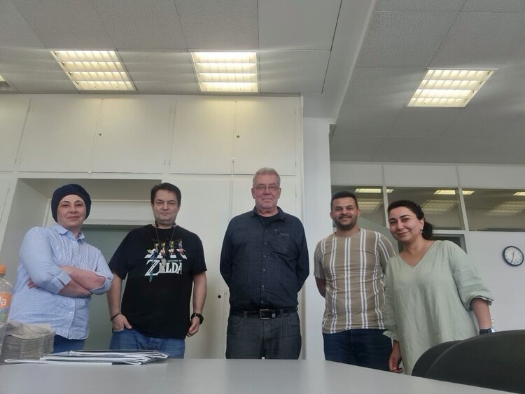 START NOW: mit Unterstützung ins Arbeitsleben In einem Klassenzimmer stehen fünf Personen für ein Gruppenfoto bereit. In der Mitte drei Männer, außen zwei Frauen. Im Hintergrund hängt eine Uhr an der Wand, auf der 12:35 Uhr angezeigt wird. Die Neonröhren an der Decke sind angeschaltet.