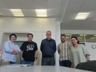Gruppenfoto In einem Klassenzimmer stehen fünf Personen für ein Gruppenfoto bereit. In der Mitte drei Männer, außen zwei Frauen. Im Hintergrund hängt eine Uhr an der Wand, auf der 12:35 Uhr angezeigt wird. Die Neonröhren an der Decke sind angeschaltet.