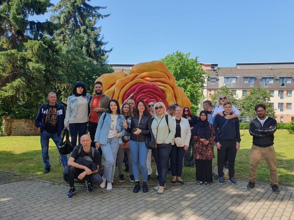 Auf den Spuren der Geschichte Zittaus Eine Gruppe von Menschen posiert für ein Foto vor einer großen, farbenfrohen Skulptur, die wie eine überdimensionale Rose aussieht. Die Gruppe steht auf einem gepflasterten Weg in einem grünen, parkähnlichen Bereich mit Bäumen und Gebäuden im Hintergrund.