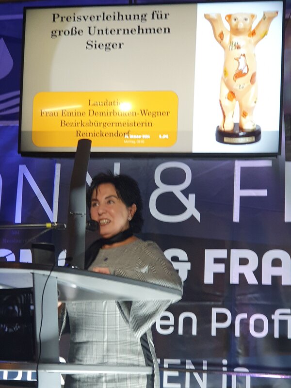 Reinickendorfer Ausbildungsbuddy geht an Netzwerkpartner der Euro-Schulen Berlin Ein Bühne mit grauem Rednerpult, an dem eine junge schwarzhaarige Frau steht, und ein Bildschirm im Hintergrund mit der Aufschrift Ausbildungsbuddy 2024.