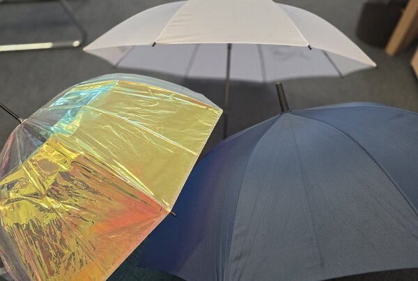 Drei verschiedenfarbige Regenschirme, die in einem Büro aufgestellt sind, geben Zeugnis von einem verregneten Sommer.