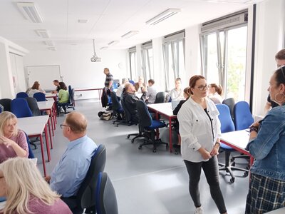 Ein heller Seminarraum mit mehreren Gruppen von Menschen, die an Tischen sitzen und diskutieren. Im Vordergrund stehen zwei Frauen im Gespräch, im Hintergrund arbeiten Teilnehmende in Kleingruppen an Aufgaben.