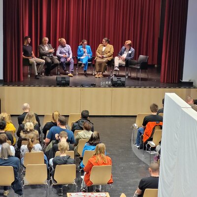 Teilnehmende sitzen im Halbkreis auf der Bühne und diskutieren, das Publikum hört zu.