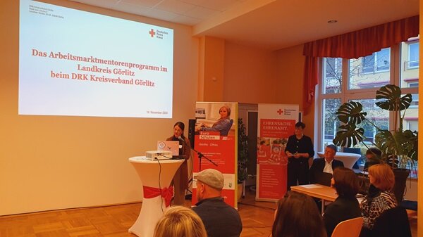 Menschen erfolgreich in Arbeit oder Ausbildung begleiten Teilnehmerin am Stehtisch spricht zu Gästen, im Hintergrund stehen Euro-Schulen und DRK Rollups.