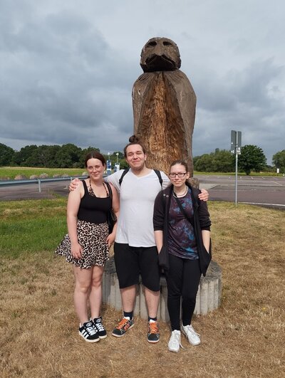 Erinnerungsfoto Die drei Auszubildenden posieren gemeinsam für ein Erinnerungsfoto vor einer hölzernen großen Skulptur, die auf einem Feld an der Ecke einer Kreuzung steht..