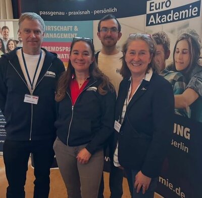 Vier Personen stehen vor einem Banner der Euro Akademie Berlin und lächeln in die Kamera. Sie tragen Namensschilder und repräsentieren die Institution im Rahmen des Zukunftstags.