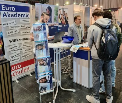 Interessierte Besucher am Messestand der Euro Akademie Zwei junge Männer mit Rucksäcken stehen vor dem Stand der Euro Akademie auf dem Karrieretag Oldenburg 2025. Ein Mitarbeiter in einem hellblauen Sakko erklärt ihnen etwas und zeigt Broschüren. Im Vordergrund ist ein Prospektständer mit Infomaterialien zu sehen. Ein weiteres Teammitglied arbeitet an einem Laptop. Der Stand ist mit Bannern gestaltet, auf denen „Euro Akademie“, "Euro-Schulen" und mehrsprachige Begrüßungen stehen.