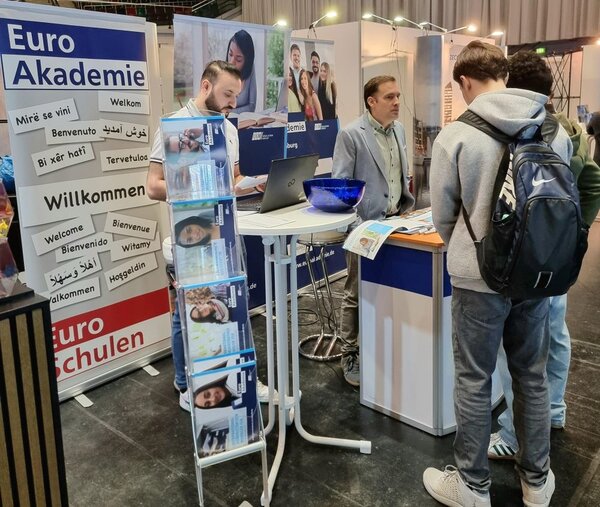 Casting für den Traumjob Zwei junge Männer mit Rucksäcken stehen vor dem Stand der Euro Akademie auf dem Karrieretag Oldenburg 2025. Ein Mitarbeiter in einem hellblauen Sakko erklärt ihnen etwas und zeigt Broschüren. Im Vordergrund ist ein Prospektständer mit Infomaterialien zu sehen. Ein weiteres Teammitglied arbeitet an einem Laptop. Der Stand ist mit Bannern gestaltet, auf denen „Euro Akademie“, "Euro-Schulen" und mehrsprachige Begrüßungen stehen.