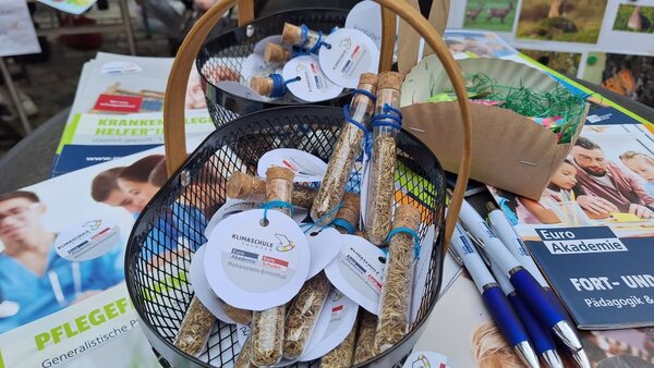 Mit Herz und Kreativität dabei Ein Korb mit kleinen Glasflaschen, die Samen enthalten, steht auf einem Tisch neben Broschüren und Stiften. Die Flaschen tragen Etiketten mit der Aufschrift 'Klimaschule'.