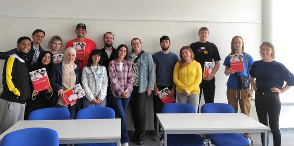 Erfolgreicher Abschluss des 100. Integrationskurses Eine Gruppe von fünfzehn jungen Erwachsenen steht lächelnd in einem Klassenraum vor einer weißen Wand. Einige halten rote Informationsmappen mit dem Titel „Fachwirt/in im Gesundheits- und Sozialwesen“. Vor der Gruppe stehen leere Tische mit blauen Stühlen.