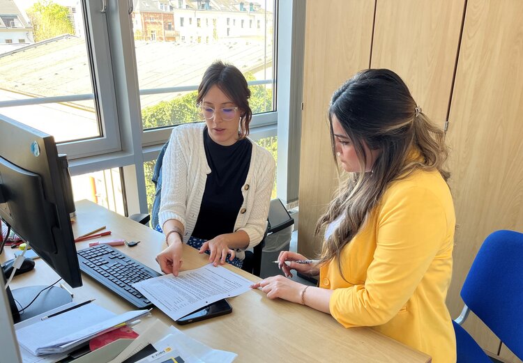 Mit individuellem Coaching zum neuen Job Zwei Frauen sitzen an einem Schreibtisch in einem Büro und besprechen gemeinsam ein Dokument; eine der Frauen zeigt mit dem Finger auf eine Textstelle, während die andere aufmerksam zuhört und einen Stift in der Hand hält.