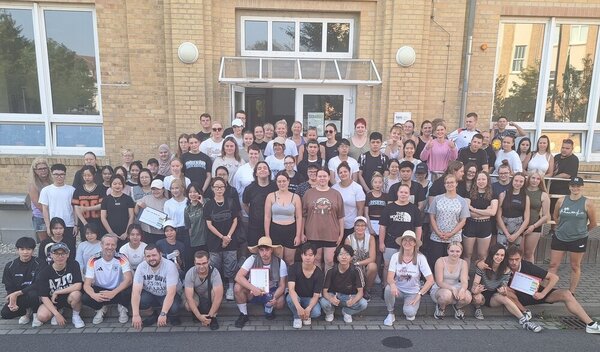 Sportfest – mal anders Ein große Gruppe von Schülerinnen, Schülern und Lehrkräften in Sportkleidung hat sich zu einem Gruppenbild vor dem Eingang des Schulgebäudes versammelt.