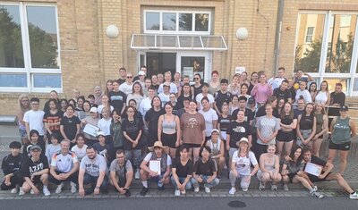 Gruppenfoto zum Sporfest Ein große Gruppe von Schülerinnen, Schülern und Lehrkräften in Sportkleidung hat sich zu einem Gruppenbild vor dem Eingang des Schulgebäudes versammelt.