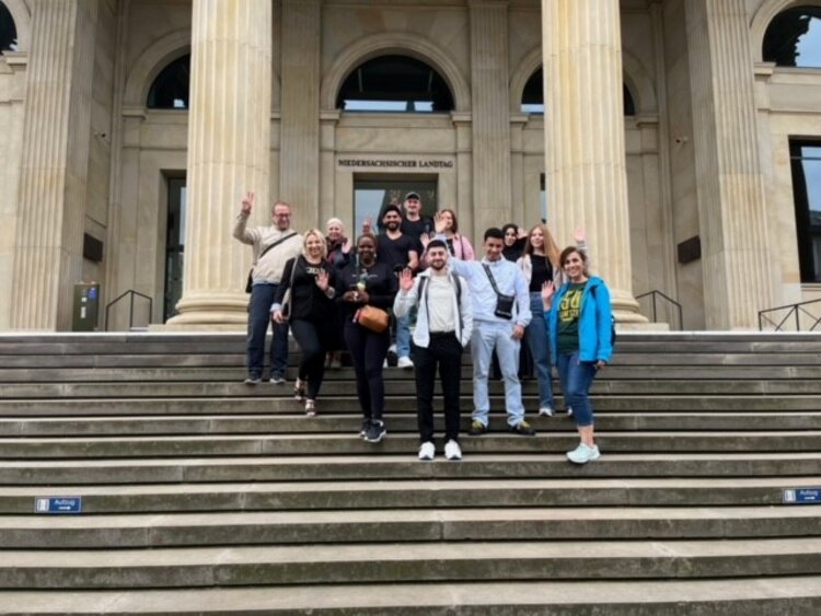 Eine Gruppe junger Menschen steht auf den Stufen vor dem niedersächsischen Landtag und winkt in die Kamera.