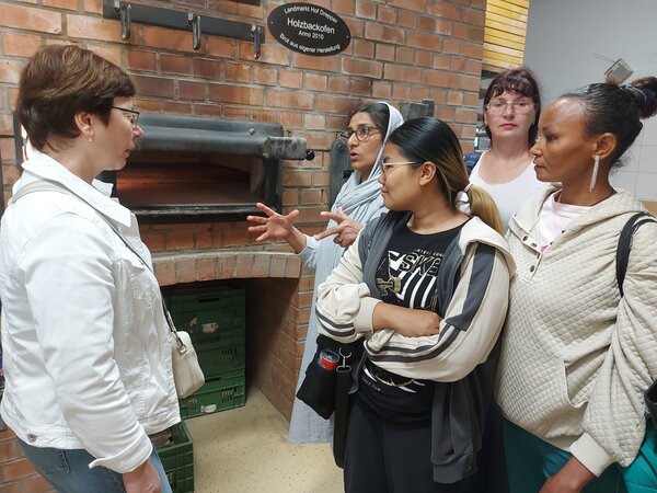 Praxis trifft Sprache - Hotel und Gastronomie Eine Frau erklärt einer kleinen Gruppe von Frauen die Funktion eines traditionellen Backofens. Die Gruppe steht vor einer Ziegelwand mit Backofenöffnung.