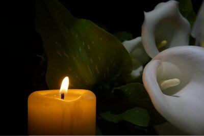 Eine Kerze scheint in der Dunkelheit, dahinter liegen zwei weiße Blumen.