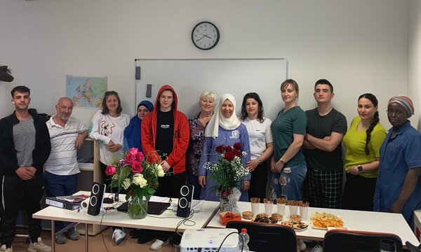 Abschiedsfeier: Feierlich und wertschätzend Eine Gruppe von Teilnehmer*innen und Lehrkräften des Integrationskurses steht lächelnd in einem Klassenraum zusammen. Auf dem Tisch vor ihnen befinden sich Blumensträuße, Lautsprecher und ein Buffet mit Snacks und Gebäck. Im Hintergrund eine Weltkarte und eine Wanduhr.