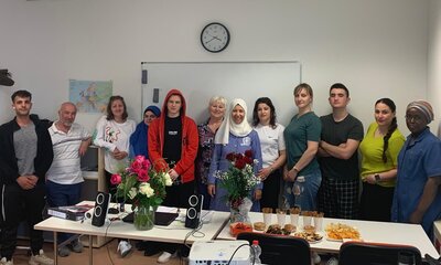Abschiedsfeier des Integrationskurses an den Euro-Schulen Gransee Eine Gruppe von Teilnehmer*innen und Lehrkräften des Integrationskurses steht lächelnd in einem Klassenraum zusammen. Auf dem Tisch vor ihnen befinden sich Blumensträuße, Lautsprecher und ein Buffet mit Snacks und Gebäck. Im Hintergrund eine Weltkarte und eine Wanduhr.