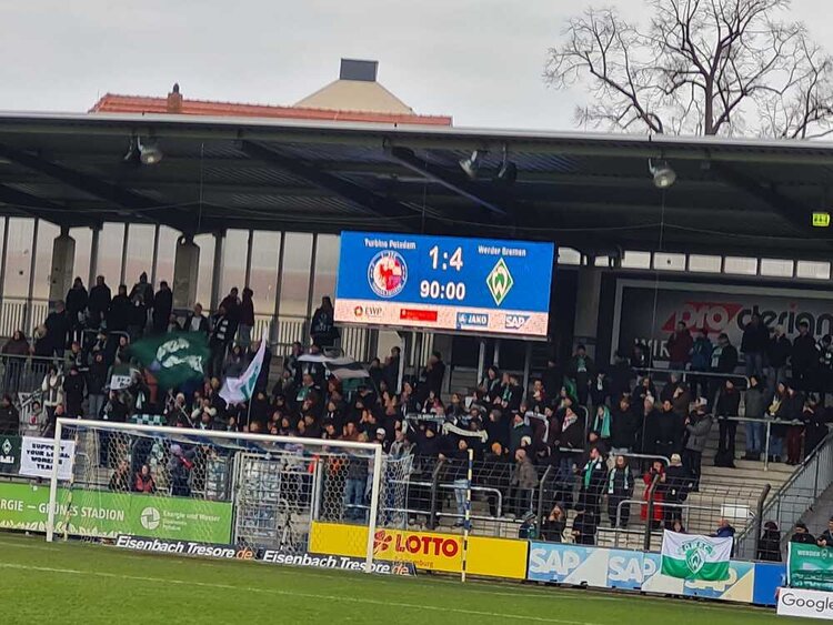 Stadionerlebnis der Extraklasse bei Turbine Potsdam Auf der Tribüne eines mittelgroßen Fußballstadions stehen Menschen mit Fahnen und Fanschals. Über ihnen thront die Anzeigetafel, die anzeigt, dass das Spiel zwischen dem Gastgeber Turbine Potsdam und Werder Bremen in der 90. Minute 1:4 steht.