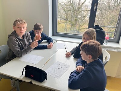Vier Schüler*innen sitzen gemeinsam an einem Tisch vor einem Fenster und arbeiten an einem Arbeitsblatt. Zwei Jungen und zwei Mädchen diskutieren, einer hält einen Bleistift hoch.