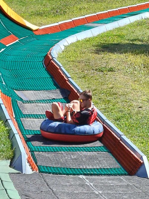 Abschlussfahrt: Erlebnispark Teichland Ein junger Mann rutscht auf einem Ring das letzte Stück einer mit Teppich ausgelegte Rodelbahn herunter. Er trägt eine reflektierende Sonnenbrille.
