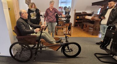 Interaktive Erfahrung mit historischem Fahrzeug Ein Mann sitzt auf einem alten dreirädrigen Fahrzeug in einem Museum, während andere Besucher zuschauen und lächeln.