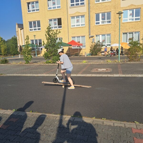 Sportfest – mal anders Auf dem Parkplatz vor dem Schulgebäude fährt eine Schülerin mit einem Roller über eine Wippe, die aus einem Stück Holz und einem langen Brett improvisiert ist. Vorne im Bild kann man die Schatten der Zuschauer auf dem Asphalt erkennen. Im Hintergrund sind entlang des Schulgebäudes Sonnenschirme und Bänke aufgestellt.