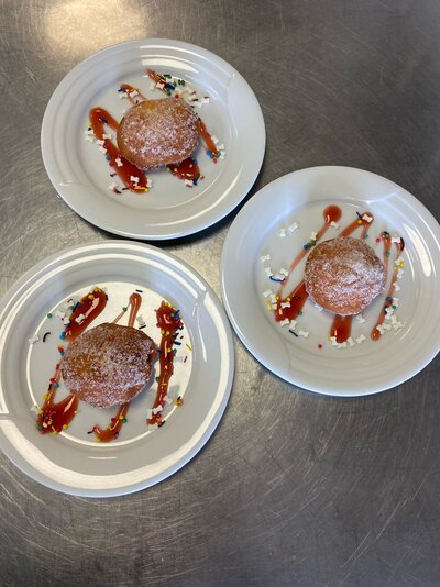 Drei weiße Teller mit jeweils einem mit Zucker bestreuten Krapfen (Berliner) sind auf einem Edelstahltisch angerichtet. Die Teller sind mit roten Soßenstreifen sowie bunten Zuckerstreuseln dekoriert.