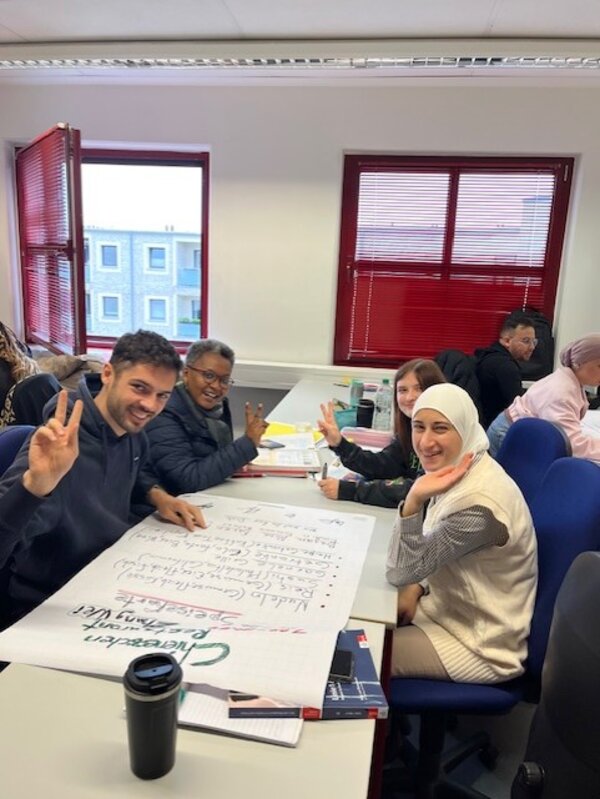 Erfolgreiche Gruppenarbeit im Berufssprachkurs Vier Teilnehmende des Berufssprachkurses sitzen an ihrer Projektarbeit und lächeln in die Kamera.