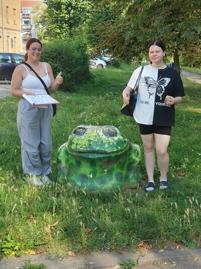 Zwei Schülerinnen stehen in einem Grünstreifen, zwischen ihnen die Skulptur eines grünen Frosches. Beide Mädchen haben Taschen umhängen und halten einen "Daumen hoch" in die Kamera. Eines der Mädchen hat eine aufgeklappte Mappe in der rechten Hand.