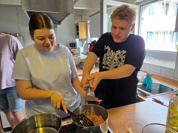 Lirum larum Löffelstiel Zwei Auszubildende stehen nebeneinander an einem Herd in einer Schulküche. Eine junge Frau rührt mit einem Pfannenwender in einem Topf mit Hackfleisch, während ein junger Mann mit beiden Händen eine Knoblauchpresse bedient. Beide wirken konzentriert und gut gelaunt.