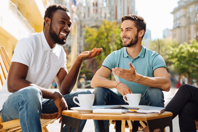 Zwei junge Männer sitzen draußen an einem kleinen Tisch mit zwei Kaffeetassen und unterhalten sich freundlich.
