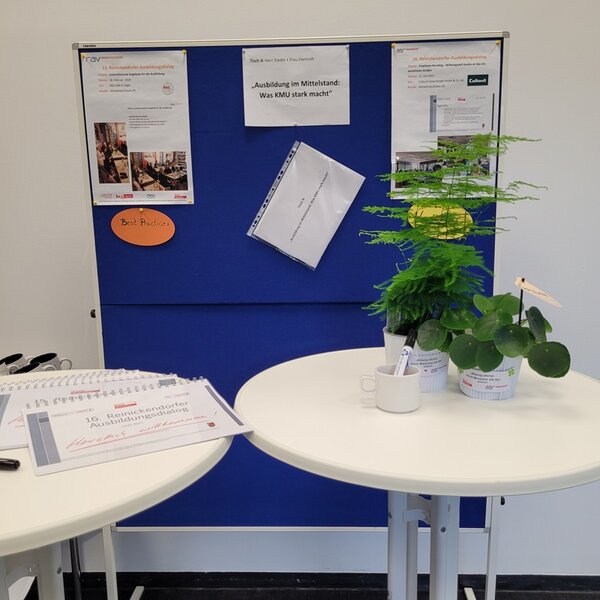 10 Jahre Reinickendorfer Ausbildungsdialog Blaues Flipchart im Hintergrund mit Informationen zum RAD, davor zwei Stehtische, auf einem davon stehen zwei Grünpflanzen.