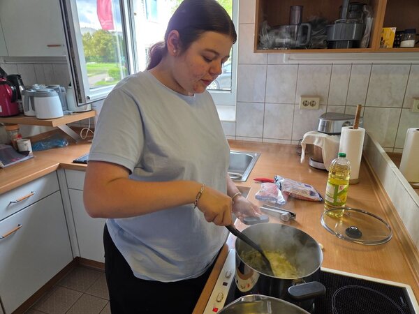 Lirum larum Löffelstiel Eine Auszubildende steht an einem Herd in einer Schulküche und rührt mit einem Löffel in einem dampfenden Topf. Im Hintergrund sind eine offene Fensterfront, Haushaltsgeräte und Zutaten zu sehen. Die Szene wirkt ruhig und fokussiert.