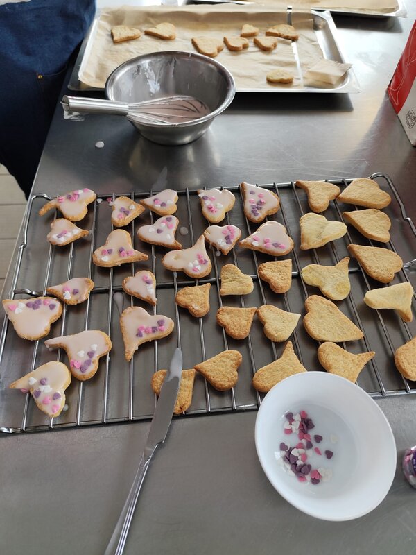 Praxisnahe Berufsorientierung: BRAFO im Strukturelement I Herzförmige Kekse werden auf einem Gitterrost glasiert und mit kleinen bunten Zuckerherzen dekoriert. Eine Schüssel mit Dekorationselementen und ein Schneebesen liegen daneben.
