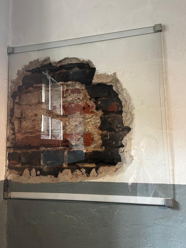 Exkursion zur Gedenkstätte Bautzen Ein Ausschnitt einer Wand mit freigelegtem Mauerwerk aus Ziegelsteinen, hinter Glas geschützt. Die Stelle wirkt wie ein restaurierter Bereich einer Gedenkstätte oder eines historischen Gebäudes.