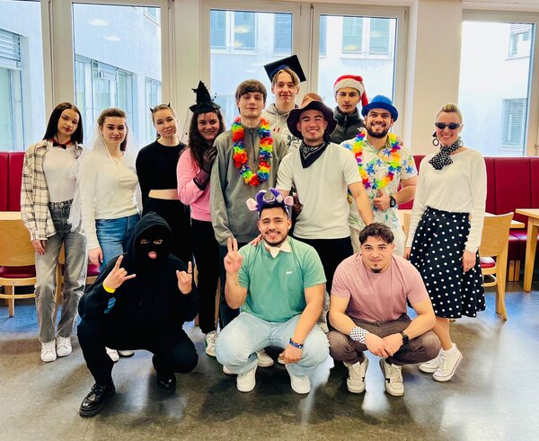 Faschingsdienstag an der Euro Akademie und den Euro-Schulen Dresden, Meißen und Riesa Eine größere Gruppe von Menschen in einem Raum mit großen Fenstern, die in verschiedenen Kostümen und Accessoires verkleidet sind. Manche tragen Hüte, Blumenketten oder Masken, während andere kreative Outfits wie ein schwarz vermummtes Gesicht oder eine Retro-Kleidung gewählt haben. Alle lächeln und posieren fröhlich für das Gruppenfoto.