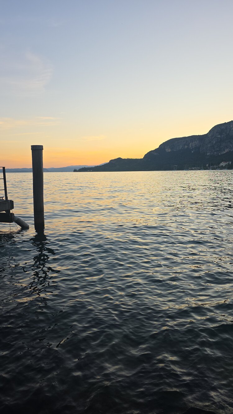 Blick auf einen Bergsee im Sonnenuntergang. An der Seite des Bildes lässt sich ein Steg erkennen. Die untergehende Sonne spiegelt sich im Wasser.