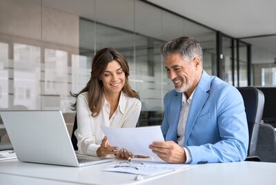 Zwei Kollegen mittleren Alters, ein Mann und eine Frau, sitzen vor einem Laptop im Büro und lesen gut gelaunt Dokumente.