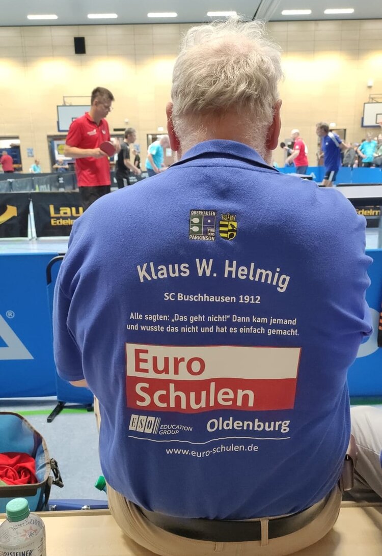 5. PingPongParkinson Quooker German Open Ein männlicher Teilnehmer an den PingPongParkinson German Open der Euro-Schulen Oldenburg sitzt auf einer Bank, mit dem Rücken zur Kamera, der rot-weiße Euro-Schulen-Aufdruck auf blauem T-Shirts ist zu sehen.