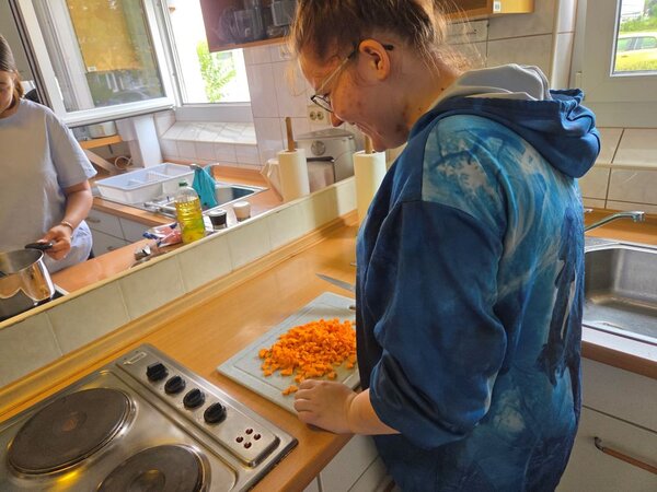 Lirum larum Löffelstiel Eine Auszubildende steht an einem Herd in einer Schulküche und schneidet mit einem Messer Karotten in kleine Stücke. Im Hintergrund sind eine offene Fensterfront, Haushaltsgeräte, Zutaten und eine weitere Auszubildende am Herd zu sehen. Die Szene wirkt ruhig und fokussiert.