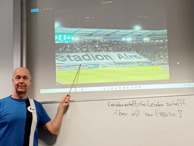 Fussball Dozent Ulrich im Arminia Bielefeld Trikot zeigt zum an die Wand projezierten Fußballspiel.