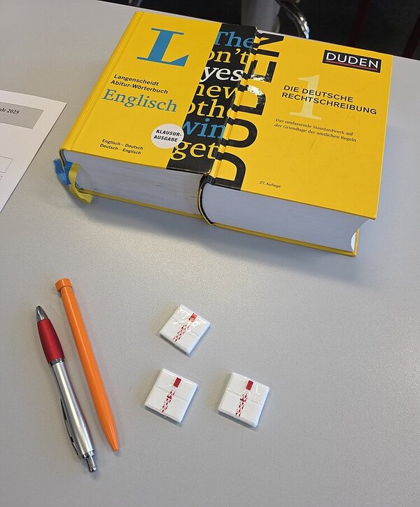 Prüfungsphase beginnt Auf einem Tisch liegen ein Kugelschreiber, ein Bleistift, Traubenzucker und ein englisches und ein deutsches Wörterbuch.