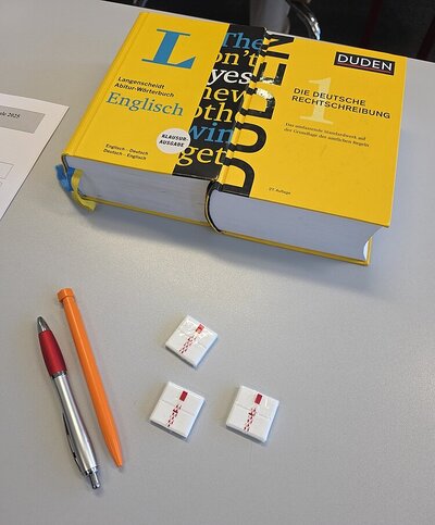 Auf einem Tisch liegen ein Kugelschreiber, ein Bleistift, Traubenzucker und ein englisches und ein deutsches Wörterbuch.