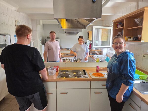 Lirum larum Löffelstiel Vier Personen befinden sich in einer Schulküche. Zwei Personen stehen im Vordergrund, eine davon hält ein Schneidebrett mit geschnittenen Karotten. Im Hintergrund sind zwei weitere Personen zu sehen, die ebenfalls in der Küche arbeiten. Die Küche ist mit verschiedenen Küchengeräten und Utensilien ausgestattet, darunter ein Herd, ein Kühlschrank, und mehrere Schränke. Es gibt auch verschiedene Küchenutensilien auf der Arbeitsplatte.