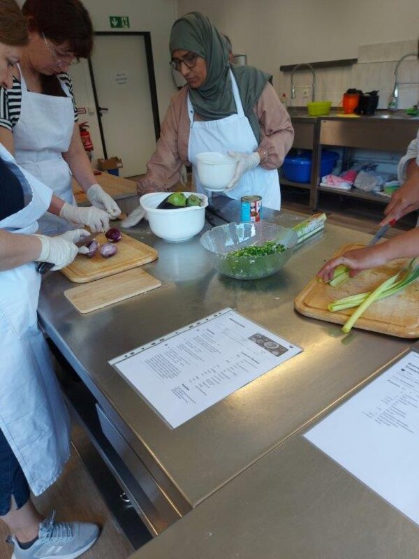 Praxis trifft Sprache - Hotel und Gastronomie Mehrere Personen mit Schürzen schneiden Gemüse auf einem Edelstahltisch, darunter rote Zwiebeln und Lauch. Auf dem Tisch liegen zudem Rezeptblätter und Schüsseln mit vorbereiteten Zutaten.