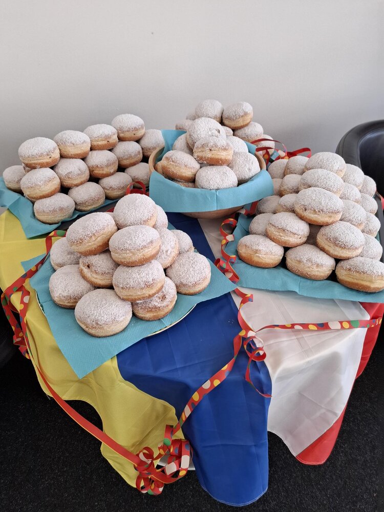 Die Euro-Schulen Mainz wünschen tolle Festnachtstage! Auf einem Tisch mit eine blau, gelb, rot und weißen Fahne liegen viele Berliner mit Puderzucker auf Tellern gestapelt.
